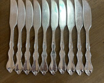 Op de afbeelding: Een set van tien zilveren botermessen op een bruin houten oppervlak. Elk mes heeft een gebogen lemmet en een decoratief handvat met een bloemmotief. De messen zijn gemaakt van glanzend, reflecterend metaal.