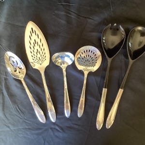 May include: A collection of six serving utensils, including a slotted spoon, a fish server, a ladle, a strainer spoon, and two serving spoons. The utensils are silver-toned with black handles on the serving spoons.