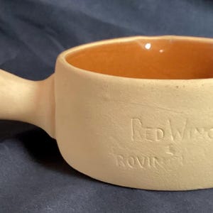 Könnte beinhalten: Eine beige Keramikschale mit Henkel. Der Innenraum ist orange. Die Worte "Red Wing" und "Rovin" sind auf der Seite eingeprägt. Die Schale hat einen Durchmesser von etwa 15 cm.