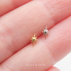 May include: Two small star-shaped studs, one in a gold tone and one in a silver tone, each featuring a four-pointed design with elongated rays. The text OLIVE & SKYE is printed in white at the bottom center of the image against the skin background.