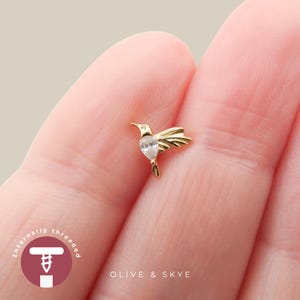 May include: A gold-toned hummingbird body jewelry stud featuring a clear, pear-shaped gemstone set in the center. The design includes a circular logo with the text Internally threaded and the brand name Olive & Skye printed below the jewelry.