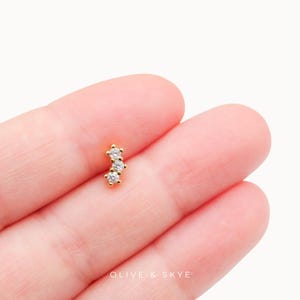 May include: A gold stud earring featuring three small, clear, round-cut stones arranged in a vertical, slightly curved line. The piece is branded with the text OLIVE & SKYE at the bottom. The design consists of a polished gold metal setting.