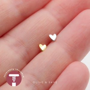 May include: Two small heart-shaped body jewellery studs, one in a polished gold tone and one in a polished silver tone. The text "Internally threaded" appears in a circular logo alongside the brand name "OLIVE & SKYE" at the bottom.