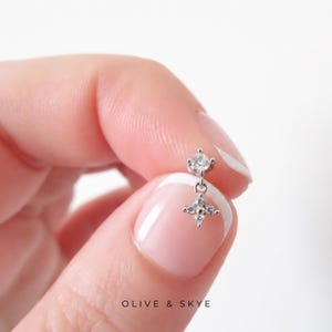 Puede incluir: Un pendiente de botón plateado con una pequeña piedra preciosa redonda y transparente en la parte superior y un dije colgante en forma de estrella de cuatro puntas con cuatro diminutas piedras transparentes. El texto OLIVE & SKYE aparece abajo.