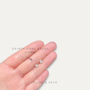 Op de afbeelding: Kleine, bloemvormige opaal oorknopjes, elk 4,5 mm groot. De oorbellen hebben een lichtblauw en wit kleurenschema. De tekst "DAINTY OPAL STUDS 4.5MM" en "OLIVE & SKYE" is ook zichtbaar.