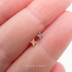 May include: Two small star-shaped stud earrings, one in gold and one in silver, each featuring a tiny central clear stone. The jewellery pieces are positioned against skin, with the text OLIVE & SKYE printed in white at the bottom of the image.