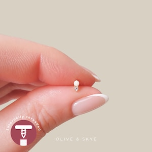 May include: A gold stud earring featuring a small round white opal stone positioned above a tiny clear gemstone. The jewellery piece is internally threaded and displays the text OLIVE & SKYE along with a screw icon in the lower corner.