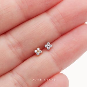 May include: Two small flower-shaped stud earrings, each featuring four clear round stones arranged in a clover pattern. One earring has a gold-toned center, while the other has a silver-toned center. The text OLIVE & SKYE is printed below.