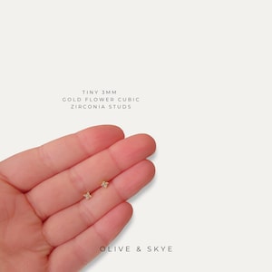 May include: Tiny gold flower cubic zirconia stud earrings. The earrings are 3mm in size and are held in a hand. The text on the image reads "TINY 3MM GOLD FLOWER CUBIC ZIRCONIA STUDS" and "OLIVE & SKYE".