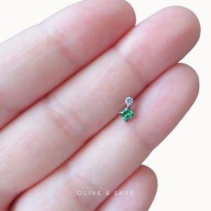 May include: A small silver-toned stud earring featuring a square-cut green gemstone positioned below a tiny, round, clear white stone. The piece is marked with the text OLIVE & SKYE at the bottom, showcasing a delicate and refined design.