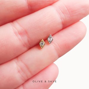 May include: Two small diamond-shaped stud earrings, one in gold tone and one in silver tone, each featuring a single clear centre stone. The text OLIVE & SKYE is printed at the bottom of the image. These jewellery pieces have a pointed geometric design.