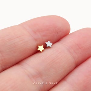 May include: Two small star-shaped stud earrings, one in a polished gold tone and one in a polished silver tone. The text OLIVE & SKYE is printed in white at the bottom of the image. The star studs are resting on a skin-toned surface.
