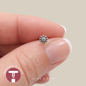 May include: A silver-toned body jewellery stud featuring a central clear gemstone surrounded by a circular arrangement of small metal beads. The piece is labelled as internally threaded and includes the brand name Olive & Skye in the graphic.