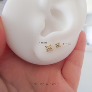 May include: A close-up of two gold-coloured flower-shaped stud earrings, 4 mm and 3 mm in size, each with clear, sparkling stones. The earrings are displayed near a white ear model. The text "OLIVE & SKYE" is at the bottom.