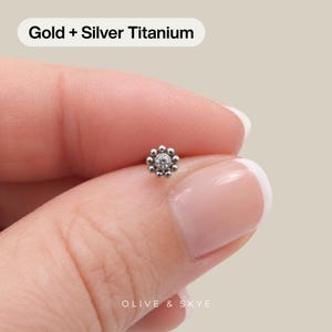 May include: A silver-toned titanium stud earring featuring a central clear round gemstone surrounded by a halo of small metallic spheres. The text Gold + Silver Titanium is visible at the top, and the brand name Olive & Skye is at the bottom.