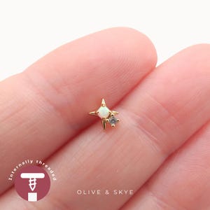 May include: A gold-toned star-shaped body jewellery stud featuring a central round white opal and a smaller clear gemstone accent. The piece includes the text Internally threaded and the brand name Olive & Skye printed on the image.