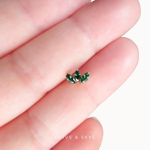 May include: A gold-toned body jewelry stud featuring three marquise-cut green gemstones arranged in a fan-like cluster. The piece is small and delicate, with the text OLIVE & SKYE printed in white at the bottom of the image frame.