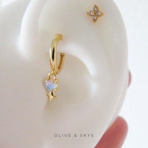May include: A gold hoop earring with a dangling charm featuring a round iridescent stone and a small four-pointed star. Above it, a separate gold stud earring shaped like a four-petaled flower with clear stones. Text reads OLIVE & SKYE.