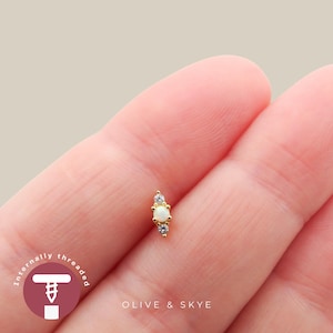 May include: A gold-toned body jewellery stud featuring a central round opal stone flanked by two smaller clear stones in a vertical arrangement. The piece includes the text "internally threaded" and "OLIVE & SKYE" printed on the image.