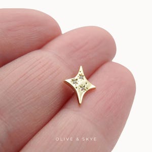 May include: A gold-toned star-shaped stud earring featuring four pointed ends and two small embedded clear stones. The surface of the earring is smooth and polished, with the text OLIVE & SKYE printed in white at the bottom of the image.