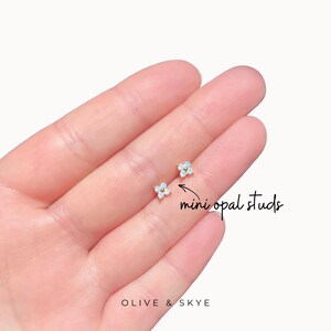 May include: A pair of mini opal stud earrings shaped like four-petaled flowers. Each earring features four small, iridescent white opal stones arranged in a clover pattern with a central gold-tone accent. The text reads mini opal studs.