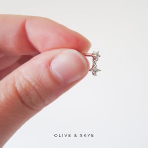 Puede incluir: Un pendiente de botón en tono plateado con un pequeño dije transparente en forma de estrella de cuatro puntas que cuelga de un poste a juego con forma de estrella. La joya se muestra sobre un fondo liso con el texto OLIVE & SKYE abajo.