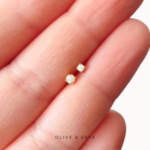 May include: A pair of small stud earrings featuring round white opals set in gold prongs. The earrings are positioned on a skin surface, and the text OLIVE & SKYE is printed in black at the bottom of the image. The design is simple and circular.
