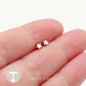 May include: Two small star-shaped stud earrings, one in a gold tone and one in a silver tone. The text "Push Pin Threadless" and "OLIVE & SKYE" is visible in the lower portion of the image, along with a circular logo featuring a stylized letter T.