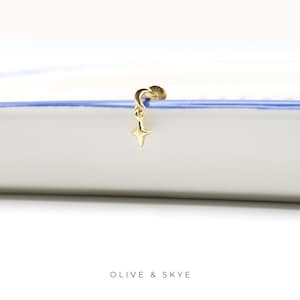 May include: A gold-toned stud earring featuring a small crescent moon charm with a dangling four-pointed star pendant. The jewelry piece has a flat back post and is displayed against a white surface with a blue edge. The text OLIVE & SKYE is visible.