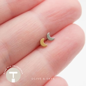 May include: Two small crescent moon stud earrings, one in a polished gold tone and one in a polished silver tone. Each moon features a smooth, solid surface and a curved shape. The text Push Pin Threadless and Olive & Skye is visible.