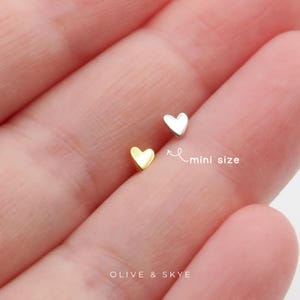 May include: Two tiny heart-shaped studs, one in a gold tone and one in a silver tone. The text "mini size" is written next to the silver heart with an arrow pointing toward it. The text "OLIVE & SKYE" is printed at the bottom of the image.