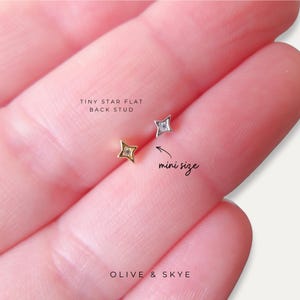 Può includere: Primo piano di due minuscoli orecchini a forma di stella. Uno è dorato, l'altro argentato, ciascuno con una piccola gemma trasparente al centro. È visibile anche il testo "TINY STAR FLAT BACK STUD" e "mini size". Il marchio "OLIVE & SKYE" è in basso.