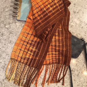 Puede incluir: Una bufanda tejida con un patrón de cuadros marrones y naranjas y flecos.