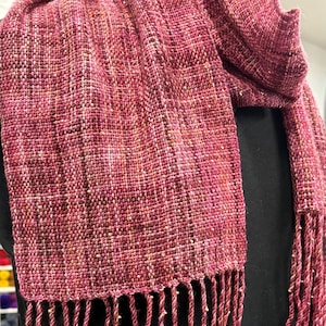 May include: A handwoven scarf in shades of pink and red with a textured weave. The scarf features a fringe at the ends and small, gold-coloured beads woven into the fabric. The scarf is draped over a dark background.