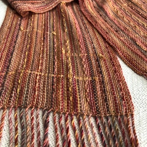 May include: A woven scarf with a fringe in shades of red, brown, and gold. The scarf is made of a fine, textured yarn and has a subtle sheen.