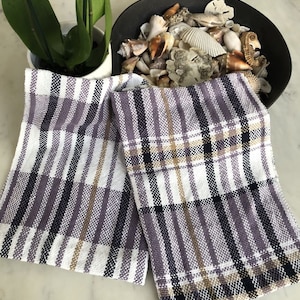 May include: Two kitchen towels with a plaid pattern in shades of purple, white, black, and tan.