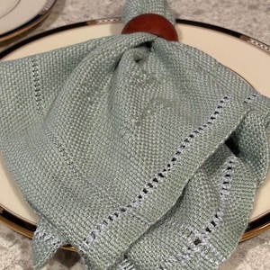 May include: A folded, light green linen napkin with a decorative border, held by a dark brown wooden napkin ring. The napkin is placed on a white plate with a gold and black rim, suggesting a formal dining setting.