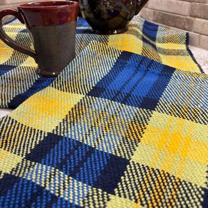 Pode incluir: Um caminho de mesa tecido com um padrão xadrez em tons de azul-marinho, amarelo brilhante e verde-claro. Uma caneca de cerâmica castanho-escura com rebordo vermelho e um bule castanho-escuro com detalhes florais estão sobre a superfície.