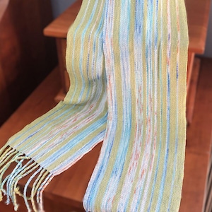 May include: A striped scarf with a green, blue, and white color scheme. The scarf has fringe on both ends.