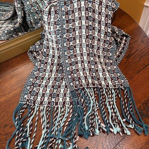 May include: A patterned scarf in teal, white, and dark brown, with a geometric design. The scarf features a fringed edge with twisted teal, white, and brown strands. It is displayed on a wooden table.