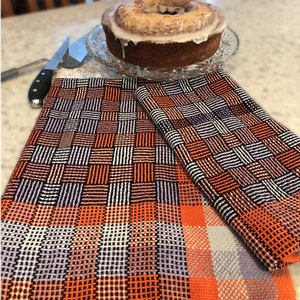 May include: Two orange and white checked kitchen towels with a black and white woven pattern. The towels are folded and placed on a white worktop with a glazed cake on a glass plate behind them.