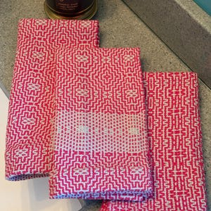 May include: Three folded pink and white patterned hand towels. The towels have a geometric design with a repeating pattern. A lit candle in a glass jar is in the background.