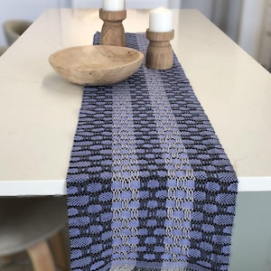 May include: A blue and gray woven table runner with a geometric pattern. The runner is on a white countertop with two wooden candle holders and a wooden bowl.