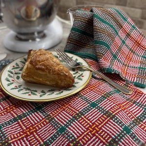 May include: A golden-brown triangular pastry sits on a white ceramic plate with a green holly leaf and red berry border. A silver fork rests on the plate. The plate is placed on a red, green, and white plaid fabric cloth with a matching folded napkin.