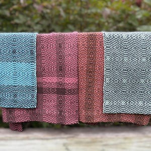May include: Four woven textiles in shades of teal, pink, and brown, each with a geometric pattern. The textiles are hanging, showcasing their intricate designs and color variations. The background is blurred, with green foliage.