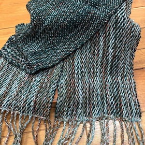 May include: A woven scarf in green, brown, and white, featuring a textured diagonal weave. The scarf has a fringed edge and is displayed on a wooden surface. The scarf appears to be made of a soft, warm material, suitable for cooler weather.