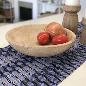Peut inclure: Un bol en bois rempli de tomates rouges et d'un oignon blanc repose sur un chemin de table tissé bleu et blanc. Deux bougeoirs en bois avec des bougies blanches sont sur le chemin de table.