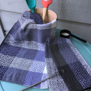 Puede incluir: Dos toallas de cocina con un patrón a cuadros negro, blanco y morado, una espátula roja con un diseño de búho, una cuchara verde y un batidor de metal sobre una mesa turquesa.