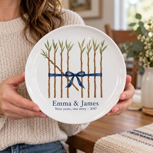 May include: A white ceramic plate featuring a watercolor illustration of nine brown willow branches tied together with a dark blue ribbon. Below the illustration, the text reads Emma & James, Nine years, one story, 2017.