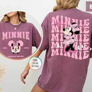 May include: A pink t-shirt with a graphic of Minnie Mouse on the back. The graphic features Minnie Mouse in a pink polka dot dress with a pink bow. The text "MINNIE" is repeated in a retro font on the back of the shirt.